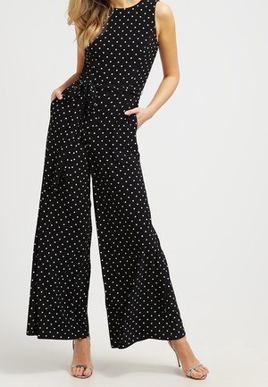 Kobieta w bezrękawnikowym czarnym kombinezonie w białe kropki, z szerokimi nogawkami, bocznymi kieszeniami i srebrnymi sandałami na wysokim obcasie, stojąca z rękami w kieszeniach.