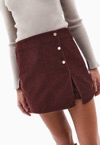 Bordo mini spódnica z miękkiego kordurowego materiału z bocznym rozcięciem, posiadająca trzy złote guziki z przodu. Tekstura jest miękka z ryflowanym detailingiem.