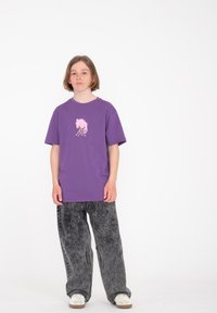 Camiseta de algodón morada con un estampado gráfico rosa, combinada con pantalones de mezclilla grises de corte holgado y acabado texturizado.