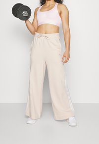 Sujetador deportivo de color rosa claro y pantalones anchos beige con franjas laterales blancas. La modelo sostiene una pesa negra. La tela parece suave y lisa.