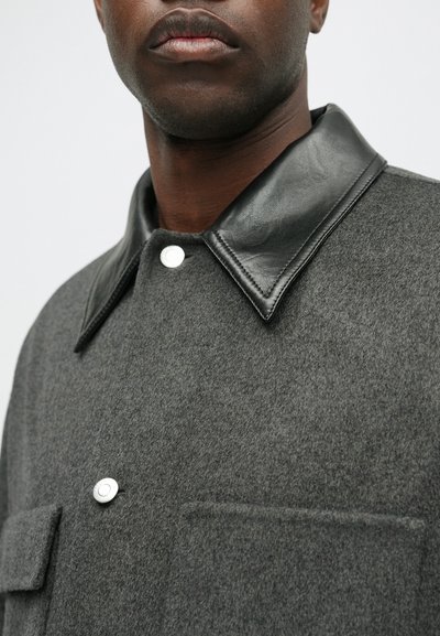 Pelēks vilnas žakets ar melnu ādas apkakli, ar priekšējo spiedpogu aizdari un krūšu kabatu, demonstrējot maigu tekstūru.