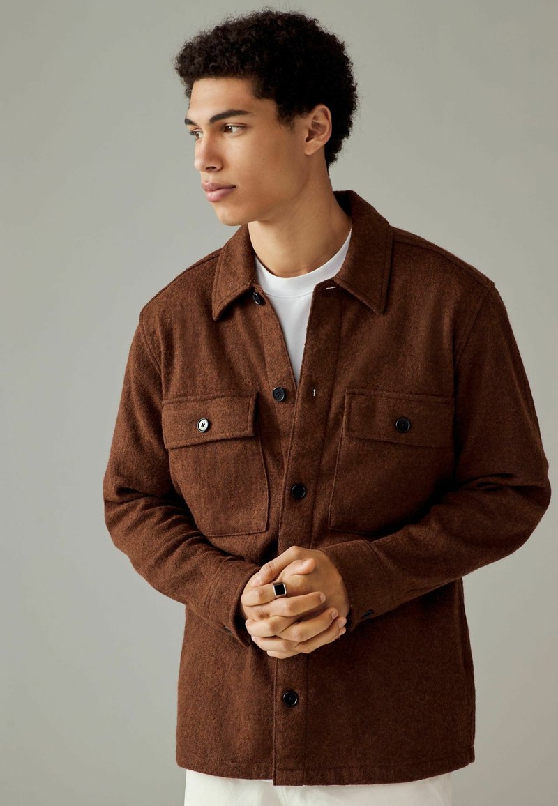 Veste en laine marron avec un col pointu, deux poches avant, boutons noirs et surface texturée, portée par-dessus un t-shirt blanc.