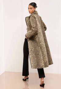 Długi beżowy płaszcz w czarne cętki, z wysokim kołnierzem, minimalnymi akcentami i luźnym krojem, w połączeniu z czarnymi spodniami i szpilkami.