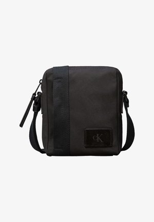 Väike must risti kehakott reguleeritava rihmaga, vertikaalne kangasdetail, tõmblukuga sulgemine ja väike logomärk esiküljel.