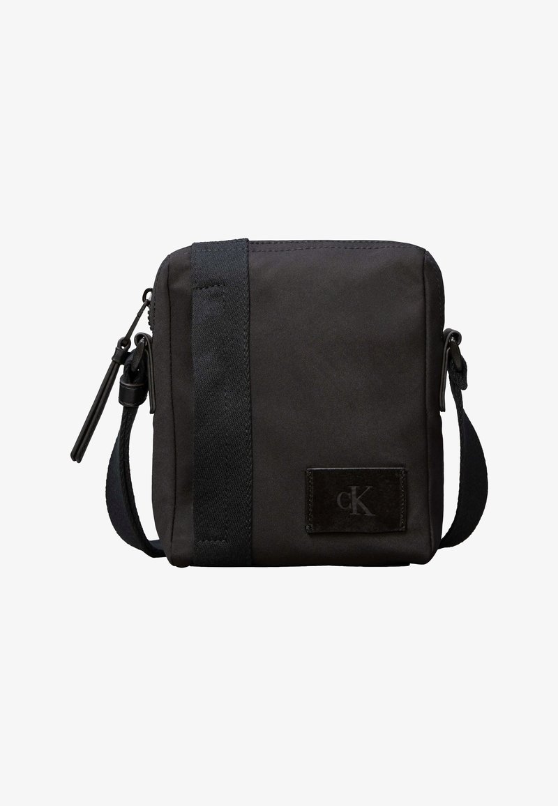 Μικρή μαύρη τσάντα ώμου με ρυθμιζόμενο λουρί, κάθετη υφασμάτινη λεπτομέρεια, κλείσιμο με φερμουάρ και μικρό λογότυπο στο μπροστινό μέρος.