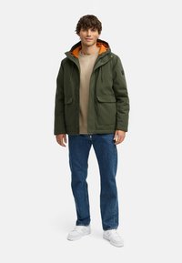 Grøn hættejakke med orange for, der har to frontlommer. Båret over en beige sweatshirts og parret med blå jeans og hvide sneakers.