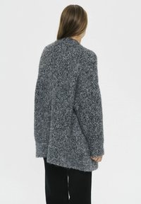 Osoba v oversized šedej pletenej kardigane a čiernych nohaviciach, stojaca chrbtom k plain bielej stene.