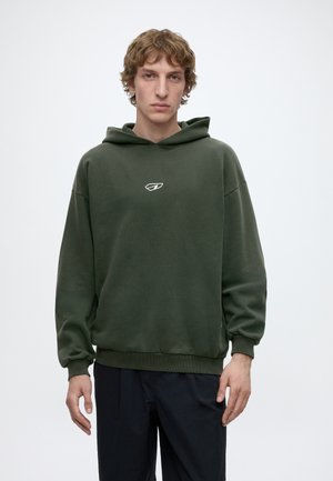 Ein junger Mann mit kurzen lockigen Haaren trägt einen dunkelgrünen Hoodie mit kleinem weißem Logo und steht vor einem schlichten hellen Hintergrund.