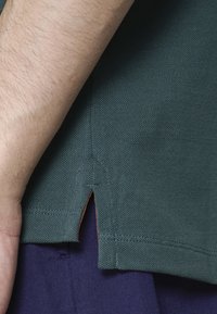 Bras reposant à côté de l'ourlet texturé d'une polo vert foncé avec une fente sur le côté, portant un pantalon bleu marine.
