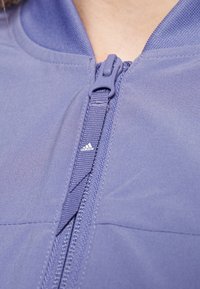 Lila sportjacka med dragkedja framtill, försedd med texturerad tyg, en platt krage och en liten vit Adidas-logga nära dragkedjan.