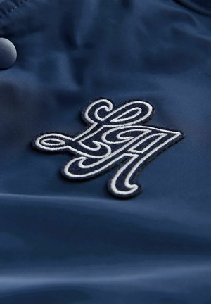 Granatowy materiał ma teksturowane, haftowane logo w białym thread, przedstawiające ozdobne litery "LA" z ciemną obwódką.