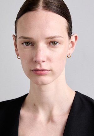 Model with straight brown hair, wearing small gold hoop earrings. She has pale skin, green eyes, and a neutral expression, dressed in a black outfit.