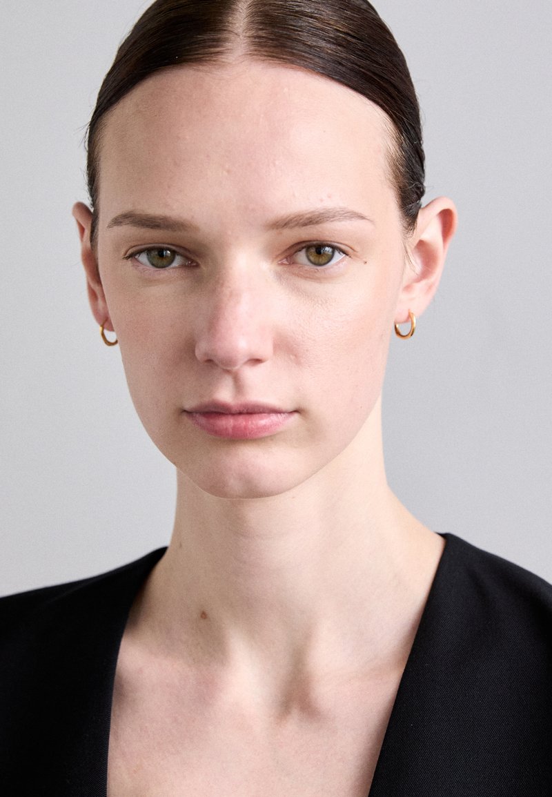 Model with straight brown hair, wearing small gold hoop earrings. She has pale skin, green eyes, and a neutral expression, dressed in a black outfit.
