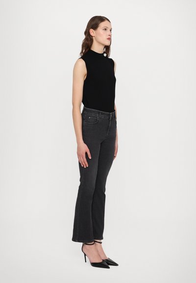Sort ærmeløs top med høj hals, parret med mørkegrå flared jeans. Modellen bærer sorte spidse hæle. Enkel, tætsiddende design.
