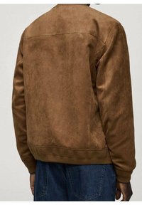 Homme portant une veste aviateur en daim marron et un jean en denim bleu, vu de dos sur un fond uni.