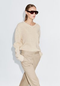 Pull en tricot beige avec un col en V, associé à un pantalon beige clair. Le modèle porte des lunettes de soleil surdimensionnées et foncées. Texture lisse, coupe décontractée.