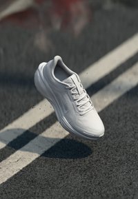 White athletic shoe with a smooth surface and mesh panels, featuring laces and a rounded toe, suspended above a dark asphalt background.