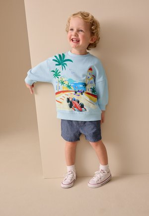 Usmievavé malé dieťa s kučeravými blond vlasmi, oblečené v modrej mikine so závodnou tematikou, tmavých šortkách, bielych ponožkách a teniskách, stojace proti béžovej stene.