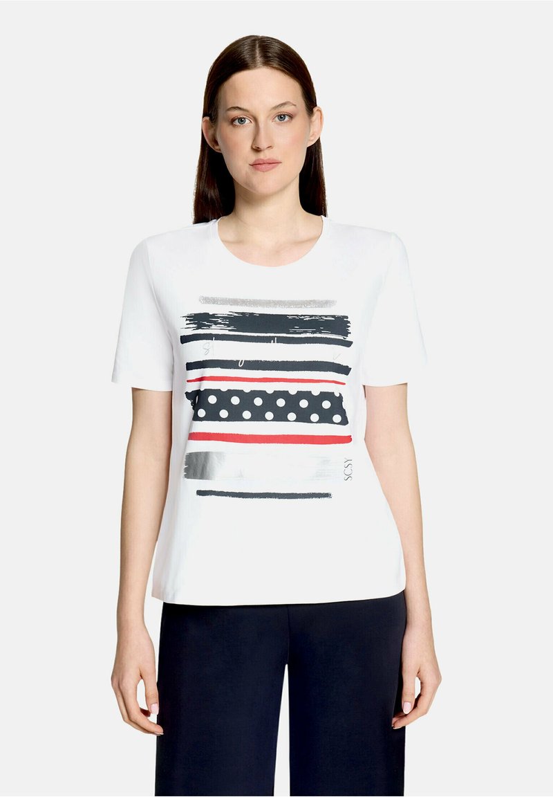 Femme portant une chemise blanche à manches courtes avec des rayures noires, rouges, argentées et des pois blancs, associée à un pantalon sombre sur un fond uni.