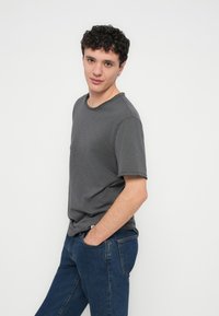 Young man with curly hair wearing a gray t-shirt and blue jeans, standing with hands in pockets, looking at the camera.