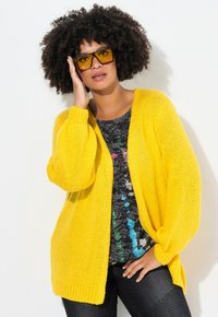 Cardigan giallo lavorato a maglia con maniche oversized, abbinato a una t-shirt nera con grafica e schizzi colorati e jeans scuri.