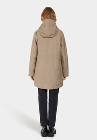 Beige wasserdichte Jacke mit Kapuze, gerader Schnitt und langen Ärmeln. Kombiniert mit schwarzen Hosen und klobigen schwarzen Stiefeln.