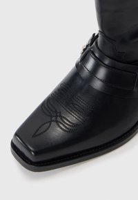 Bota de cuero negra hasta el tobillo con punta cuadrada, costuras decorativas y un accesorio de hebilla lateral. Textura suave y diseño elegante.