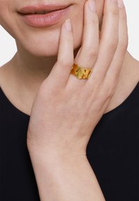 Anello placcato oro con un design ondulato, che riflette la luce. Una mano è posizionata vicino al viso, evidenziando la forma e la texture dell'anello.