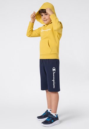 Geel hoodie met een voorvak en wit logo, gecombineerd met marineblauwe shorts met een wit logo. Zwarte en blauwe sneakers.