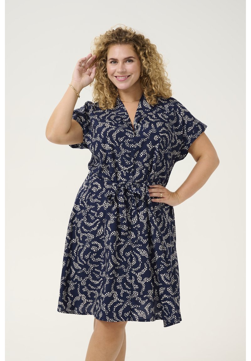 Dunkelblaues Kleid mit einem weißen abstrakten Muster, kurzen Ärmeln, Kragen und einem Bindegürtel in der Taille. Aus leichtem Stoff gefertigt.