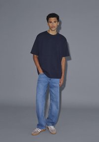Ung mand står med den ene hånd i lommen iført sort oversized t-shirt, blå jeans og grå sneakers mod en ensfarvet grå baggrund.
