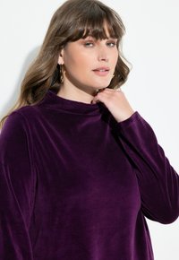 Lila, samtige Bluse mit hohem Kragen, langen Ärmeln und einer weichen Textur. Das Model berührt ihren Halsbereich und zeigt die Passform des Kleidungsstücks.