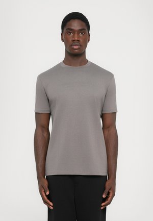Camiseta gris de manga corta en tejido suave con cuello redondo, que presenta un sutil logotipo en la parte delantera. Combinada con pantalones negros.