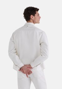 Chaqueta de mezclilla blanca con mangas largas, cuello con botones y detalles de costura visibles. El modelo muestra la chaqueta desde la espalda, con las manos entrelazadas.