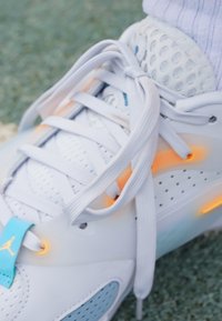 Weißer Sportschuh aus glattem Material mit Netzakzenten und orangefarbenem LED-Detail an der Sohle. Verfügt über flache Schnürsenkel und ein blaues Logo-Täfelchen.