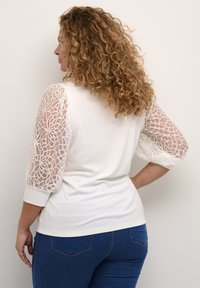 Top blanco con mangas abullonadas de encaje, con un patrón floral texturizado. La espalda es lisa, combinado con jeans azules.