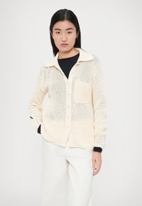 Κρεμ χρωματιστή πλεκτή ζακέτα με γιακά, κούμπωμα με κουμπιά, μπροστινή τσέπη και ανοιχτή υφή ύφανσης. Φορεμένη πάνω από μαύρο τοπ και λευκό παντελόνι.