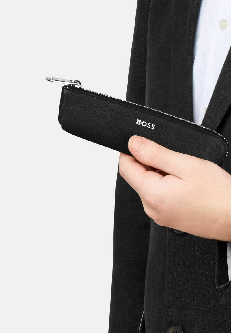 Main tenant une pochette zippée en cuir noir texturé avec le mot BOSS embossé en lettres argentées.