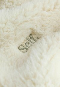 Biały, puszysty materiał o miękkiej teksturze; na jasnym tle znajduje się haft "Self." w zielonym kolorze.