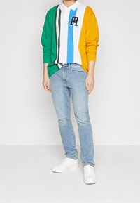 Bunter Langarm-Polo mit grünen, gelben, blauen und weißen Streifen, dazu ein schwarzes Logo, getragen mit hellblauen Jeans und weißen Sneakers.
