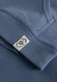 Modrá mikina s jemnou texturou, žebrovanými manžetami a bílou značkovou nášivkou na lemu. Detailní pohled ukazuje designové detaily.
