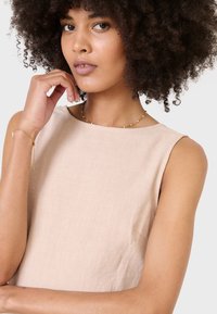 Top sin mangas color beige con un acabado texturizado, que presenta un escote redondo y suaves caídas. Un collar con detalles dorados agrega un toque especial.