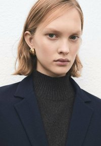 Młoda osoba z krótkimi blond włosami, nosząca złote kolczyki koła, ciemny prążkowany golf i granatową marynarkę na jasnym tle.