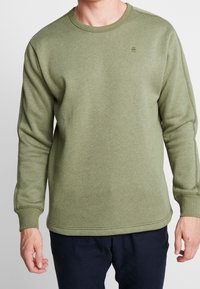 Hombre vestido con un suéter de cuello redondo verde oliva y pantalones oscuros, de pie con los brazos relajados a los lados contra un fondo blanco.