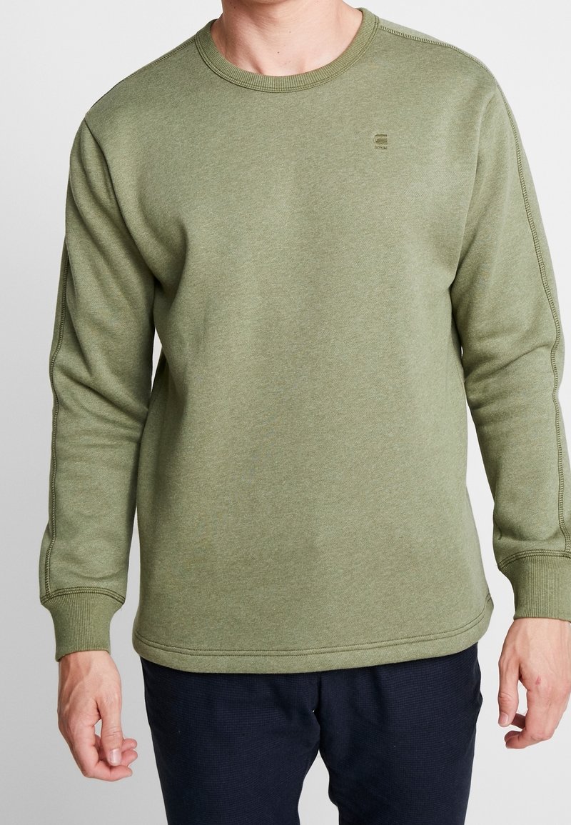 Hombre vestido con un suéter de cuello redondo verde oliva y pantalones oscuros, de pie con los brazos relajados a los lados contra un fondo blanco.