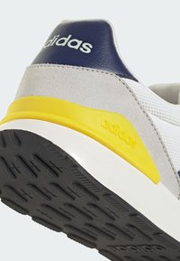 Gros plan sur le talon d'une basket Adidas présentant un mesh blanc, un daim gris, un détail bleu marine avec logo, une bande jaune et une semelle noire texturée.