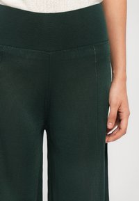 Temnozeleni rebrasti hlače z visokim pasom in stranskimi razporki. Tkanina ima teksturiran videz, kar ustvarja strukturiran izgled.