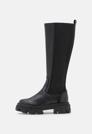 Botte noire hauteur genou avec semelle crantée épaisse, avant en cuir lisse, dos en tissu extensible et languette de maintien en haut.