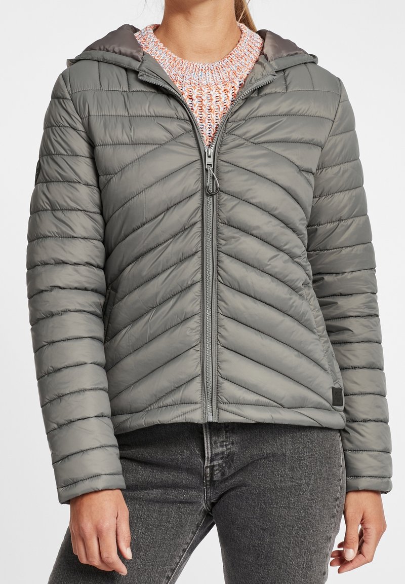 Femme portant une veste matelassée verte à capuche par-dessus un pull en maille rose et un jean gris foncé, debout devant un fond uni.