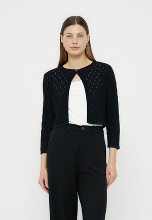 Žena s dlhými rovnými vlasmi v čiernom krátkom otvorenom pletenom cardigane cez biele tričko a čierne nohavice s vysokým pásom, stojaca na bielom pozadí.